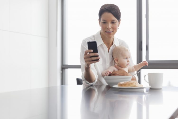 Woman holding baby in kitchen using mobile phone