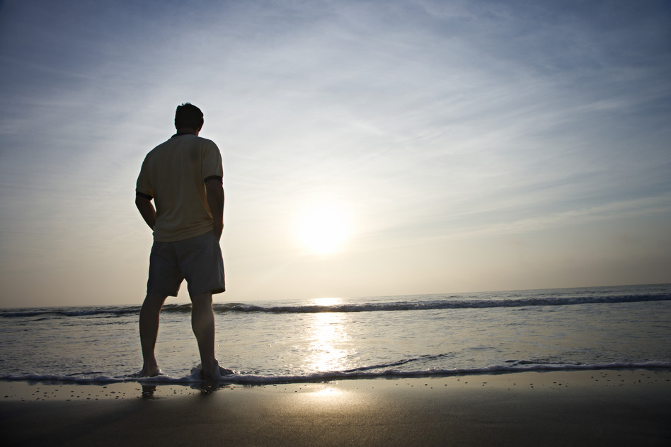 Caucasian mid-adult man standing alone on beach looking at ocean at sunrise.